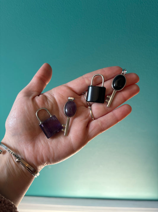 Hand holding silver lock and key pendants with black and purple stones on teal background