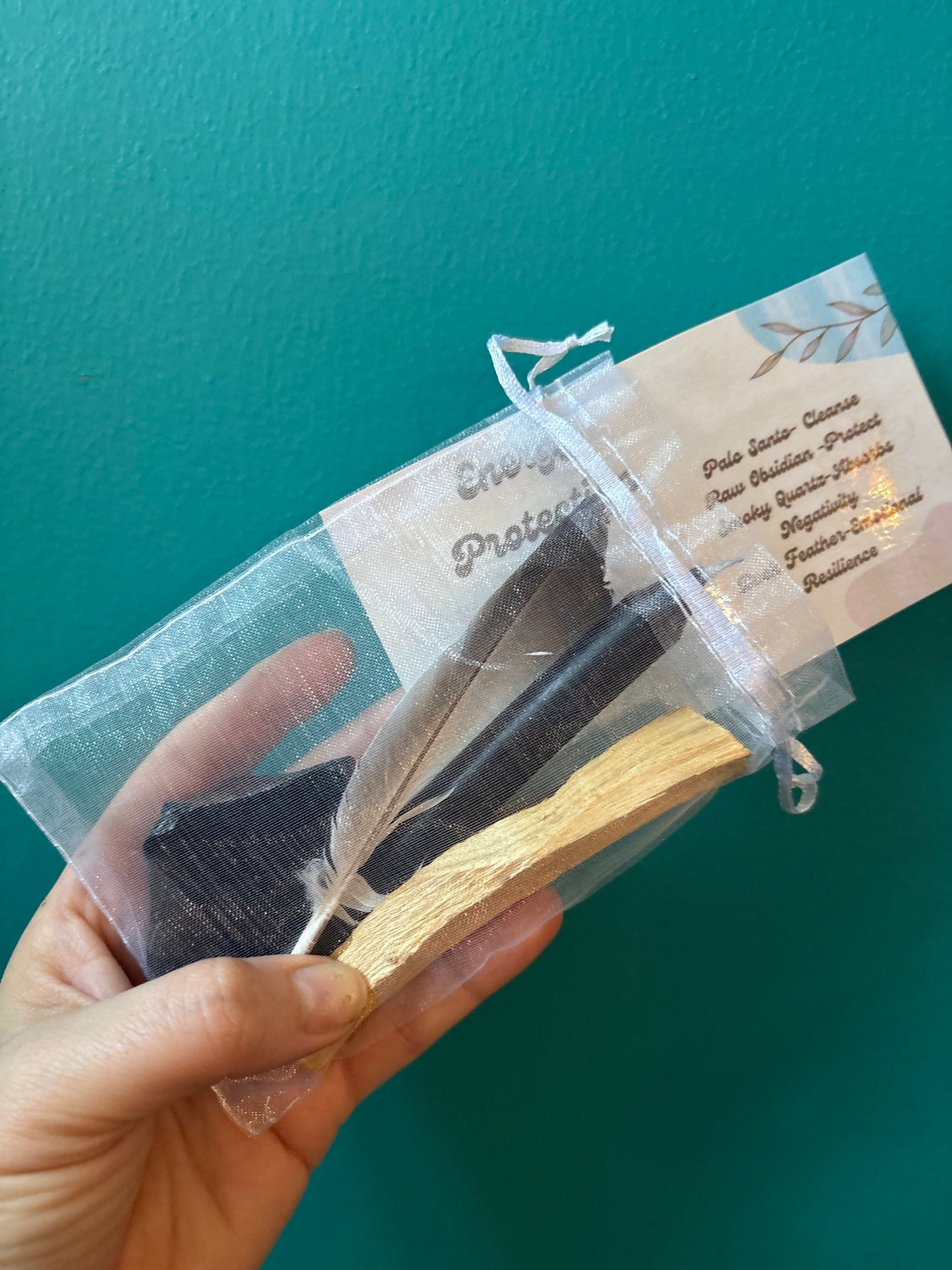 Hand holding Energy Protection Kit with cleansing tools in a transparent pouch against teal background