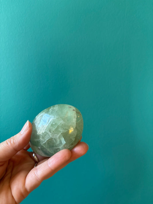 Hand holding a green Fluorite palm stone promoting mental clarity and focus against teal background