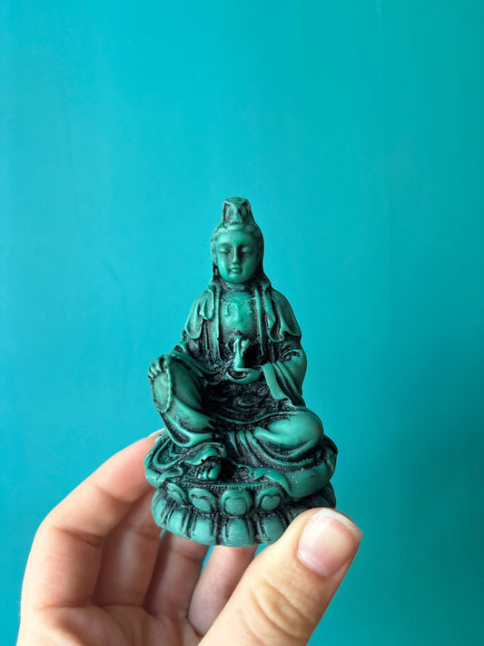 Hand holding a vibrant turquoise Quan Ying statue symbolizing compassion and wisdom against a matching turquoise background