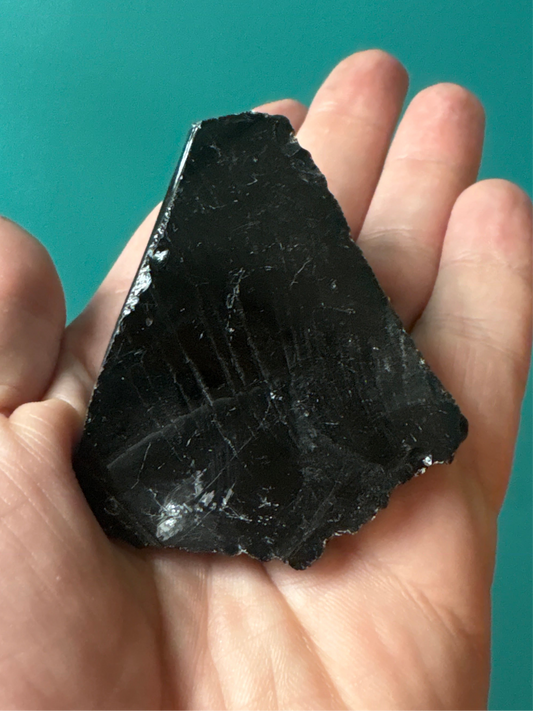 Hand holding a smooth, dark black raw obsidian stone with sharp edges against a teal background
