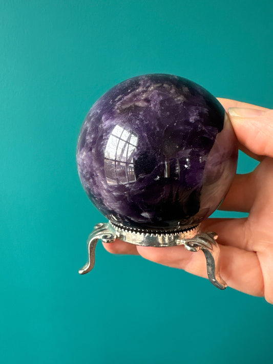 Ornate metal sphere stand holding a polished amethyst crystal ball against a teal background