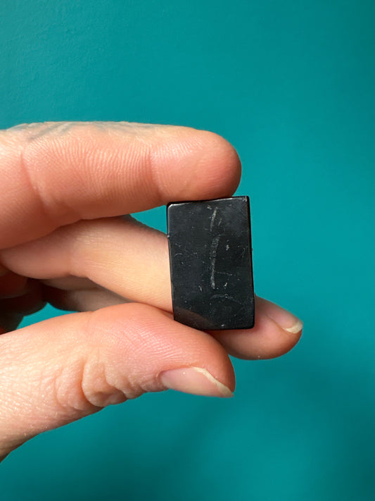 Hand holding black rectangular Shungite sticker for EMF protection against teal background