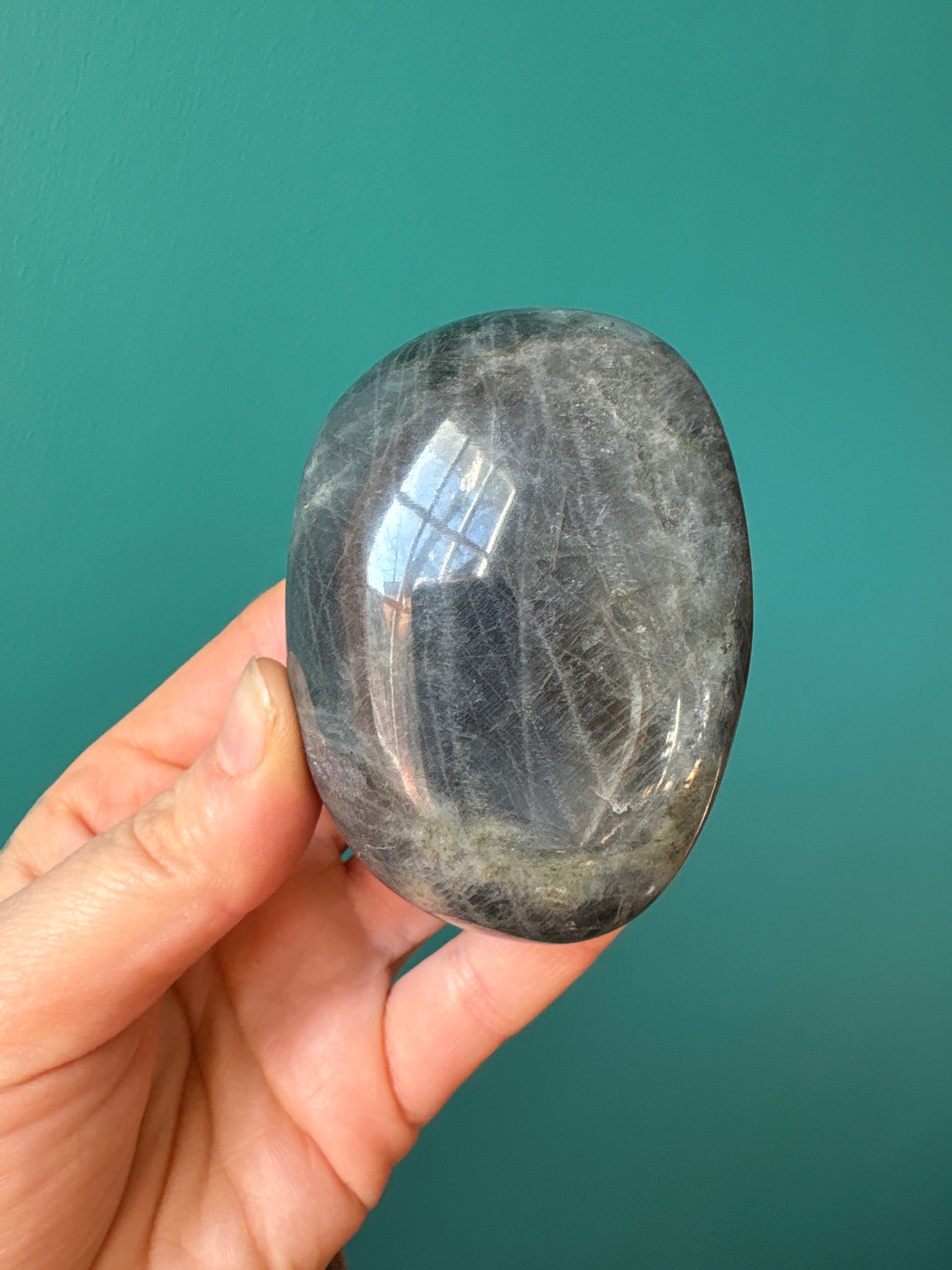 Hand holding polished Labradorite pebble palm stone with a smooth reflective surface against a teal background
