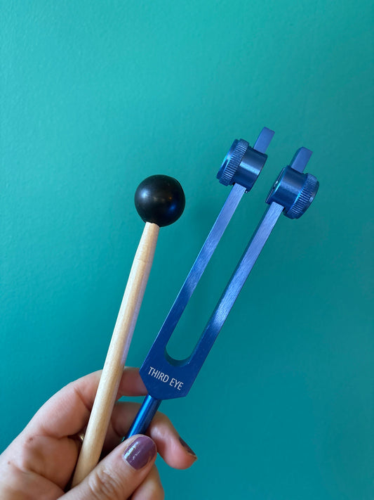 Hand holding blue Third Eye Chakra tuning fork with wooden mallet against teal background