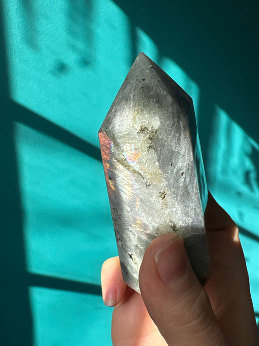 Hand holding a polished pink labradorite tower crystal with blue background and sunlight shadows