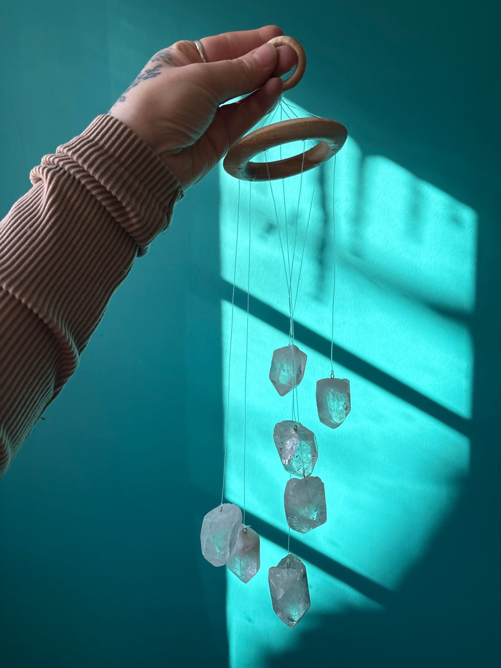 Hand holding a clear quartz wind chime with wooden rings against a teal wall background