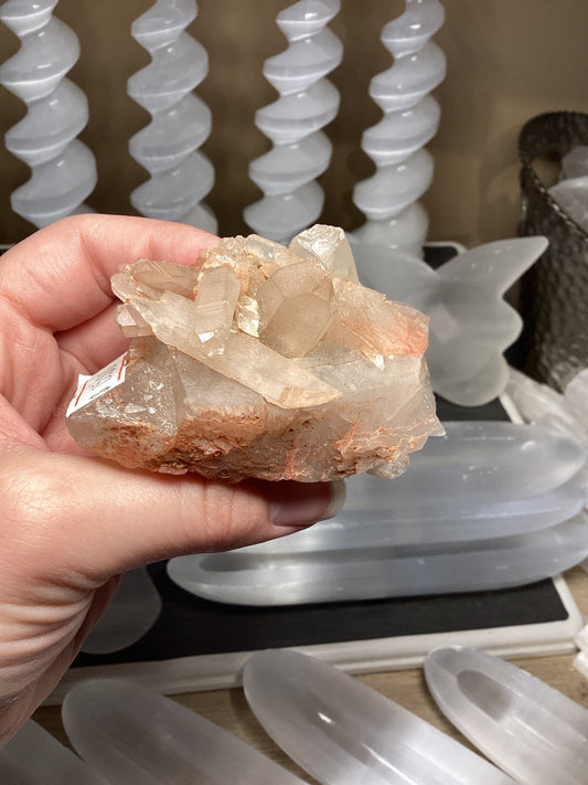 Hand holding a fire quartz cluster with reddish-orange hues and intricate crystal formations against a background of white spiral and butterfly-shaped crystal decorations