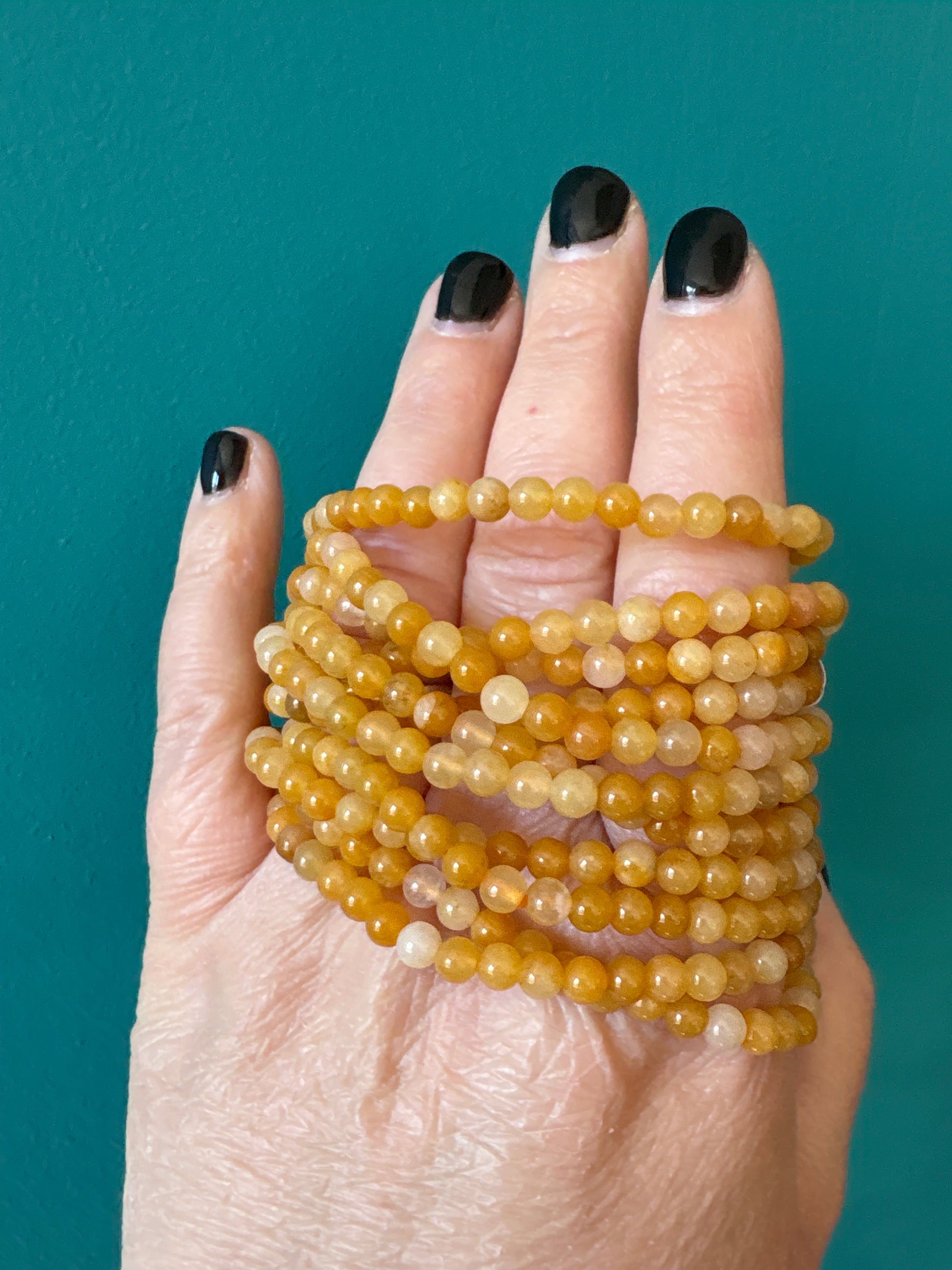 Yellow Jade 4mm bead bracelet wrapped around hand with black nail polish on teal background