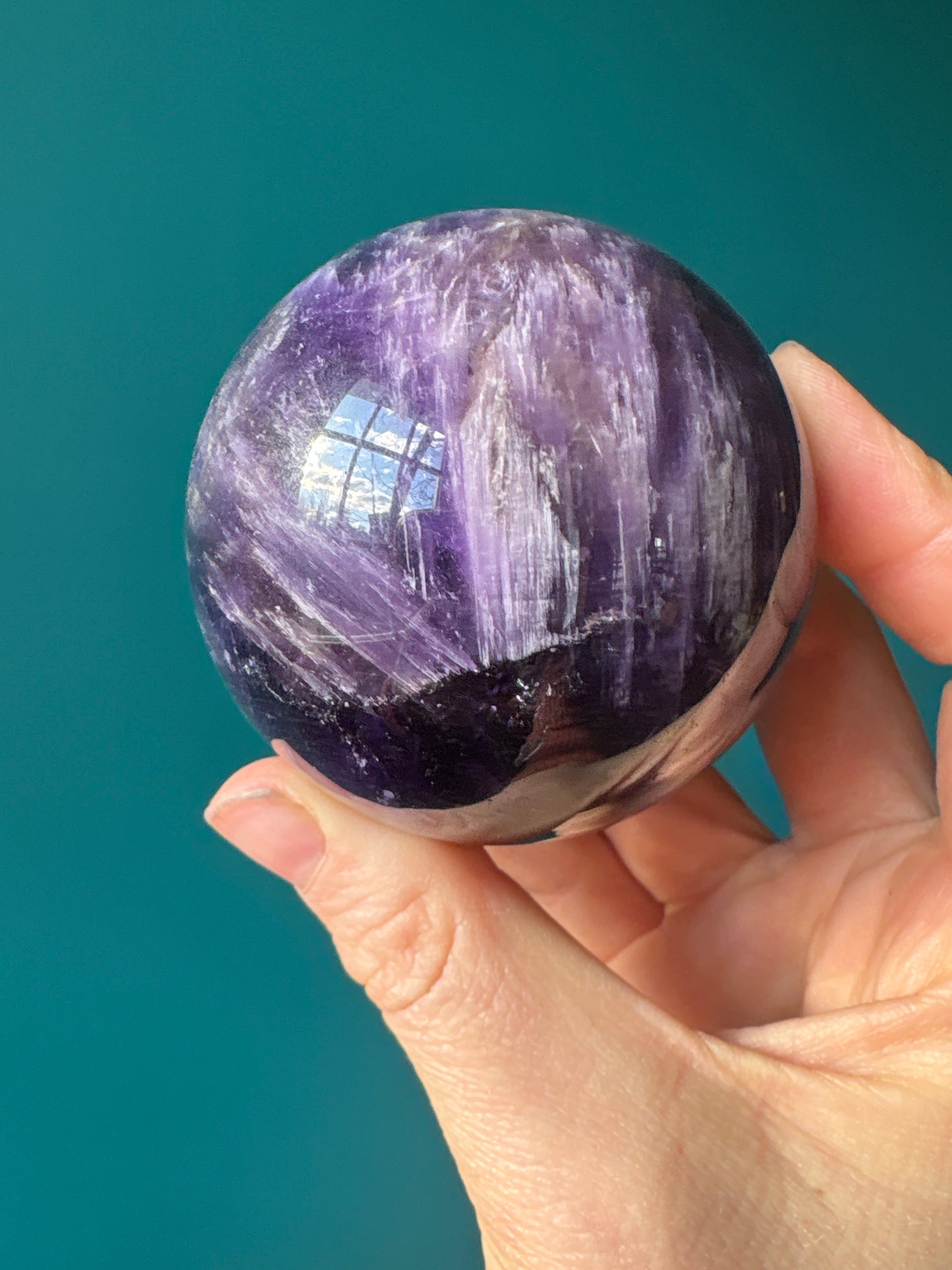 Hand holding a polished purple amethyst sphere with natural mineral patterns against a teal background