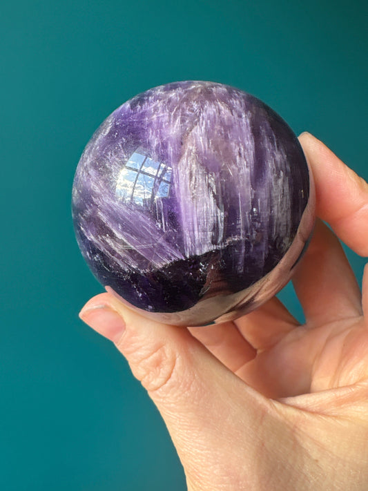 Hand holding a polished purple amethyst sphere with natural mineral patterns against a teal background