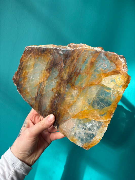 Hand holding a large hematoid quartz slab with golden healer and fire quartz colors against a teal background