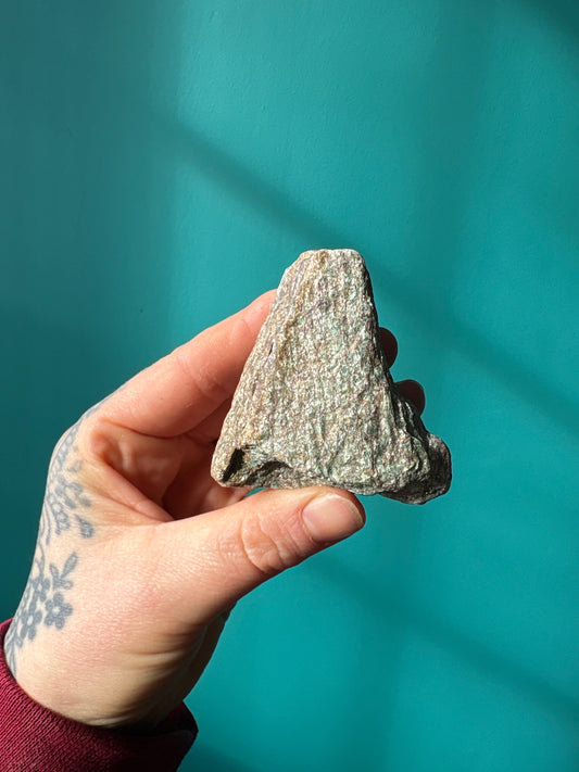 Raw Ruby Zoisite stone held against teal background showing green and red natural crystal texture