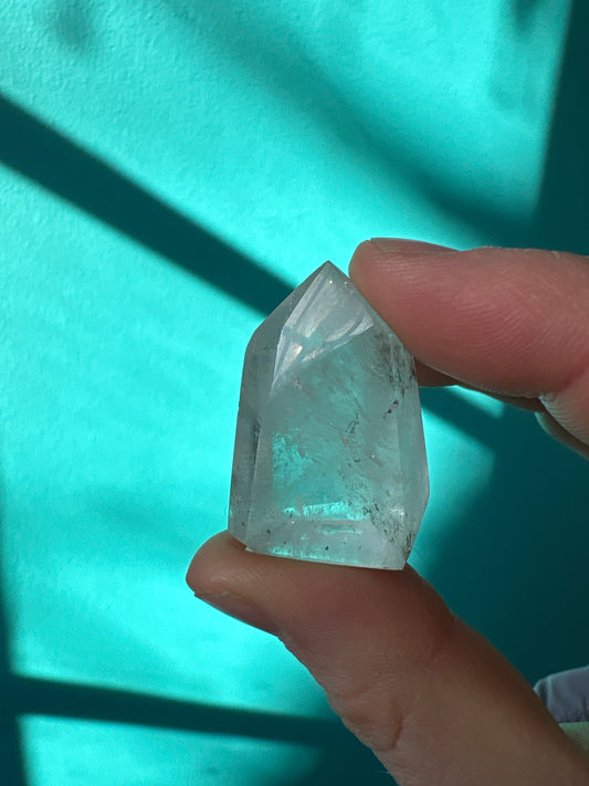 Small clear quartz point crystal held between fingers with teal background