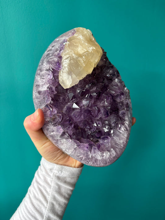 Hand holding an amethyst cluster with quartz, druzy, and gypsum crystals against a teal background