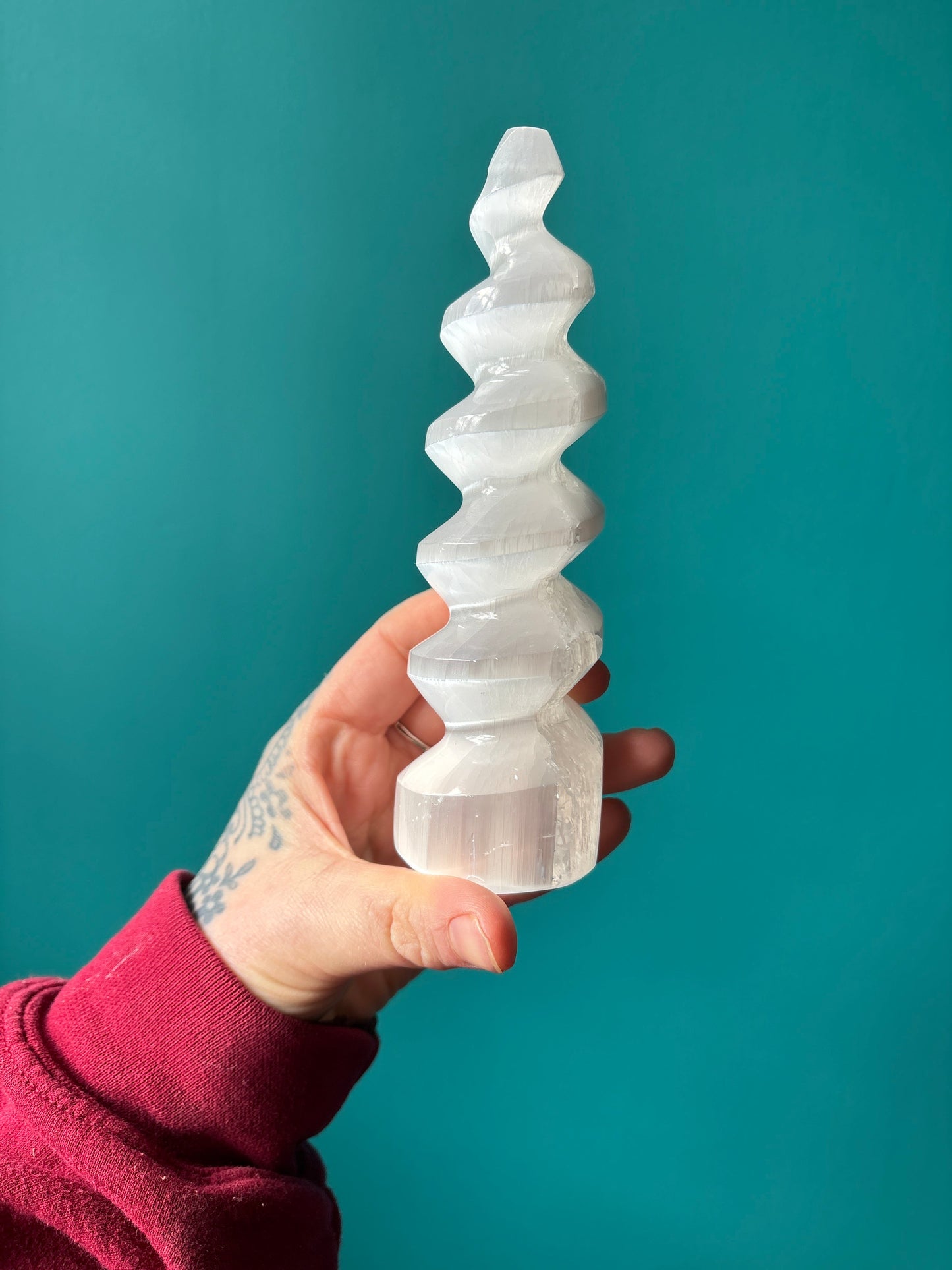 Hand holding a white spiral-shaped Unicorn Horn Selenite crystal against a teal background