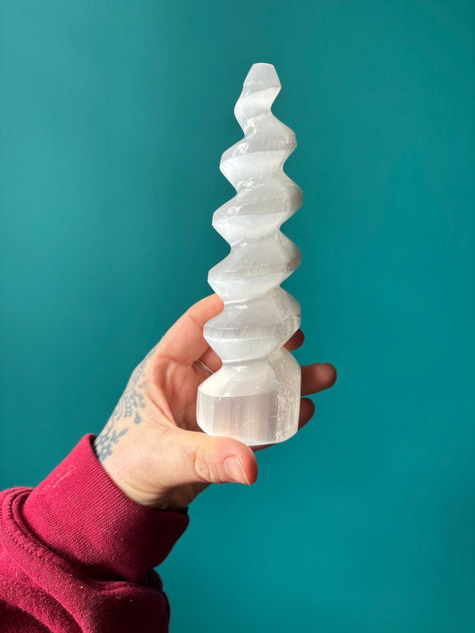 Hand holding a white spiral-shaped Unicorn Horn Selenite crystal against a teal background