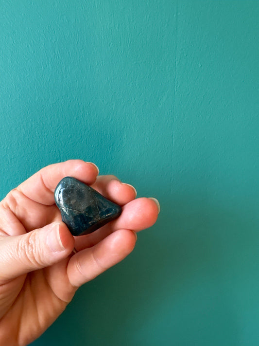 Hand holding a polished tumble blue apatite stone against a teal background, symbolizing throat chakra healing and communication enhancement.