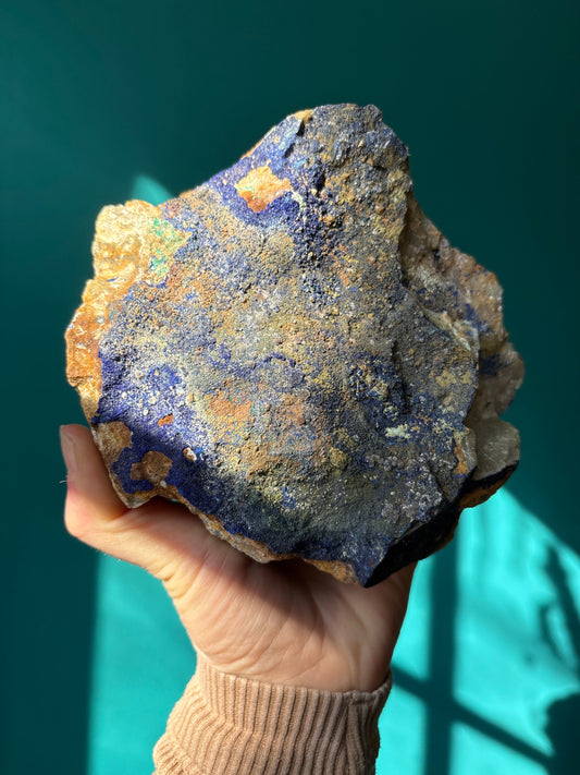 Hand holding a raw Azurite gemstone with blue and brown mineral textures against a teal background