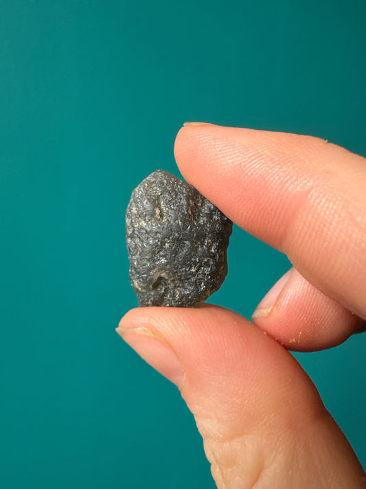Close-up of a hand holding a small tumble Agni Manitite tektite meteorite impact stone against a teal background