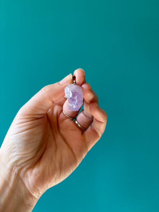 Hand holding a raw amethyst flower pendant on teal background