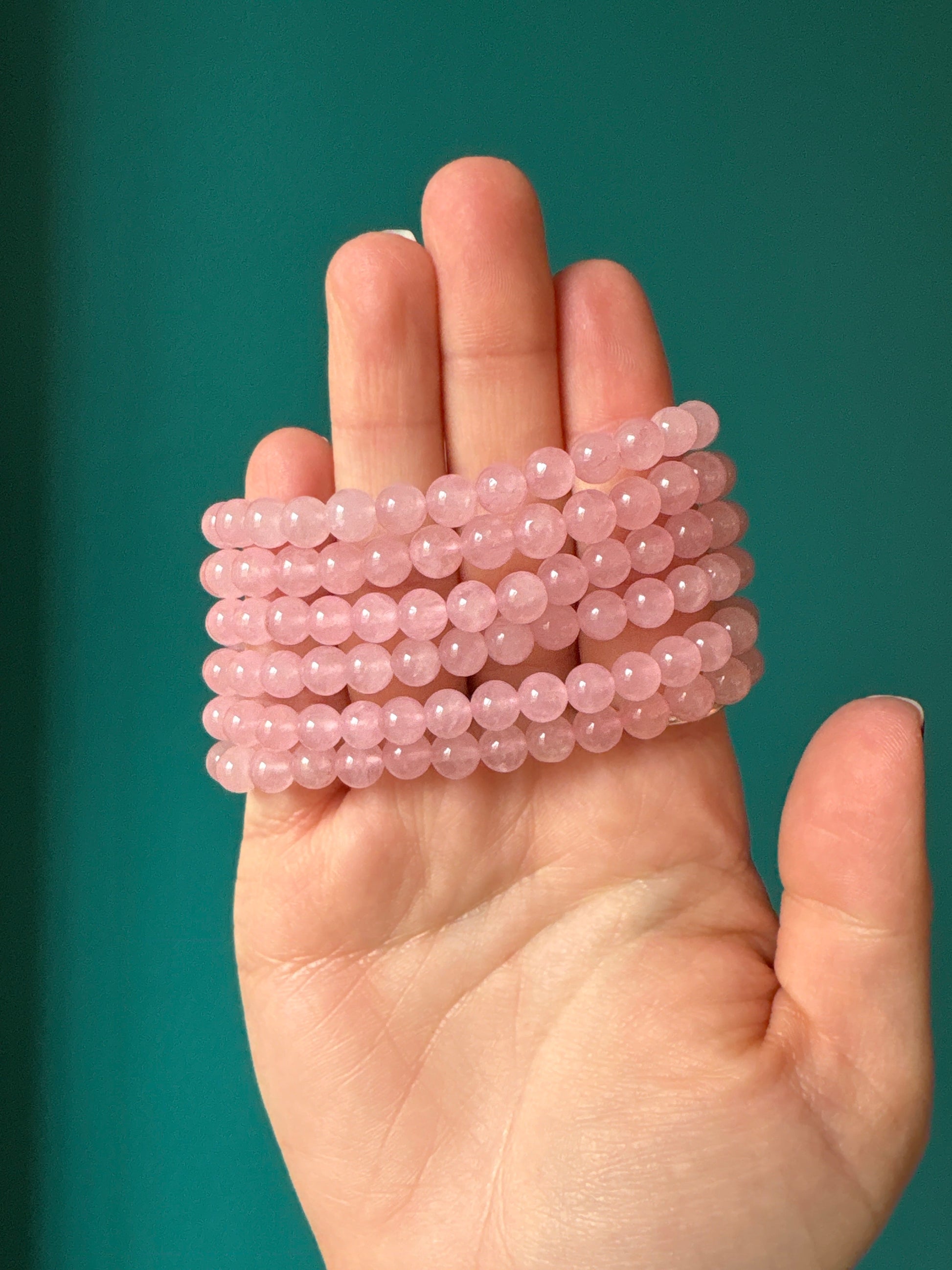 Close-up of a hand holding multiple strands of 6 mm Rose Quartz bead bracelets against a green background