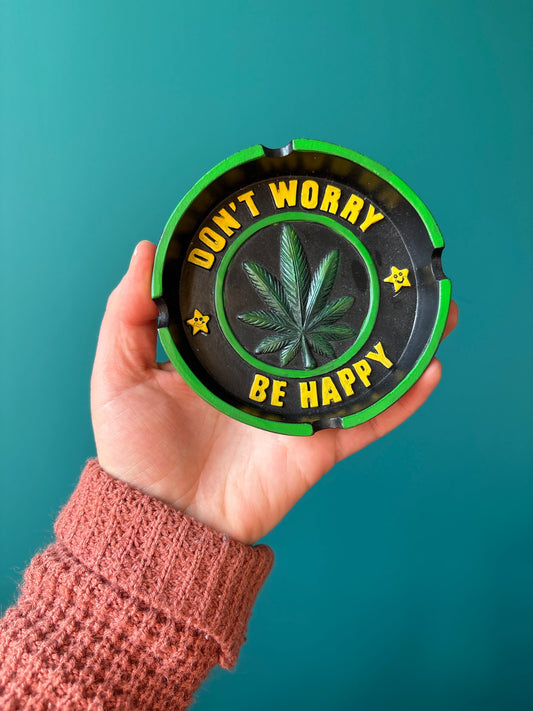 hand holding a green and black worry ashtray with a cannabis leaf and the text "Don't worry be happy"