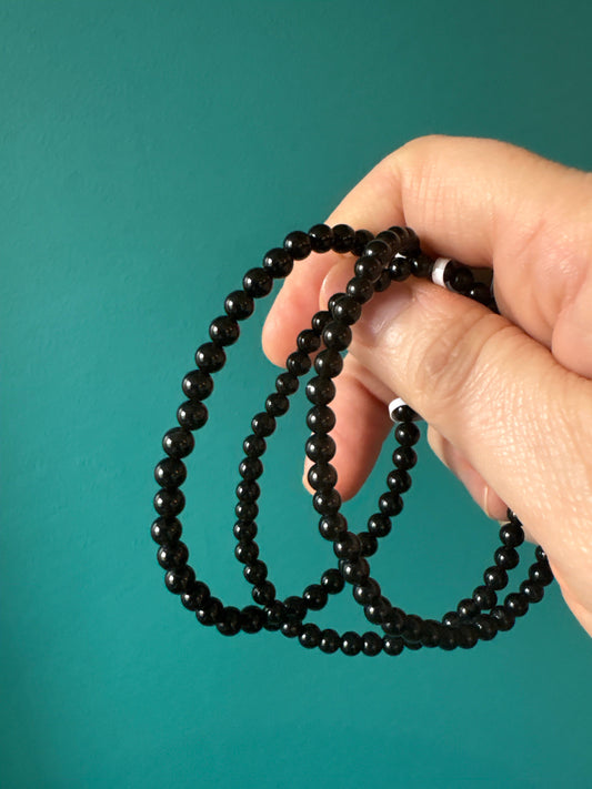 Hand holding black obsidian 4mm bead bracelet against teal background