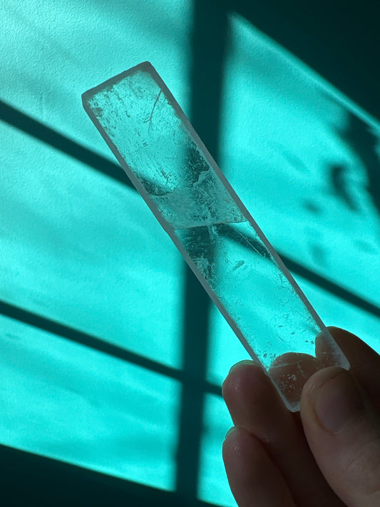 Clear selenite stick held against a teal background with light and shadow patterns