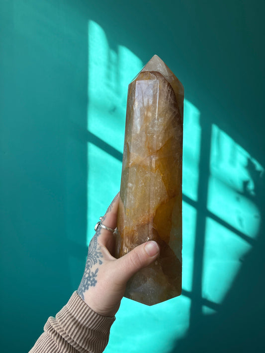 Hand holding a large Tower Golden Healer crystal with teal background and sunlight shadows