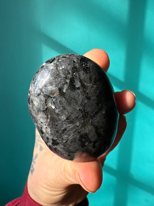 Hand holding polished Larvakite palm stone with a blue-green background for meditation and healing