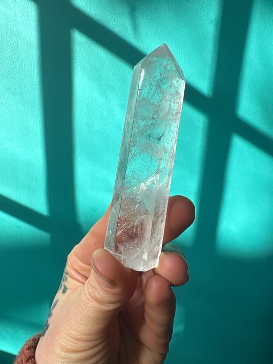 Hand holding a 3-4 inch clear quartz tower crystal against a turquoise background