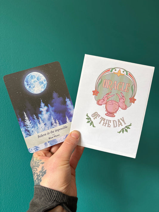 Hand holding an Oracle of the Day card reading featuring a Blue Moon card with the message "Believe in the impossible" and the Oracle of the Day booklet against a teal background