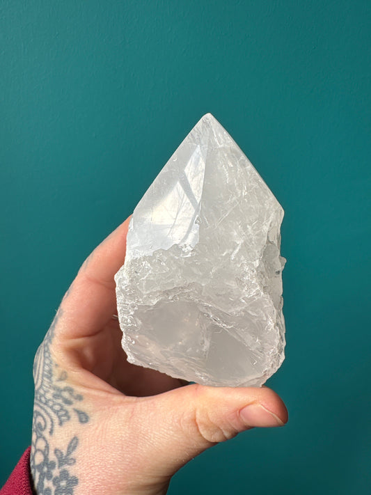 Clear quartz half polished point crystal held by hand with tattoo against teal background