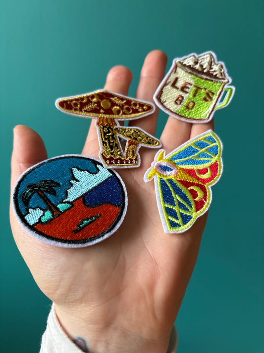 Hand holding colorful embroidered patches including a mushroom, coffee mug with "Let's Go" text, palm tree landscape, and multicolored moth designs