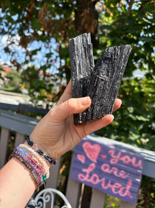 Hand holding two black tourmaline raw wands with a blurred outdoor background and a "You are Loved" sign in the distance