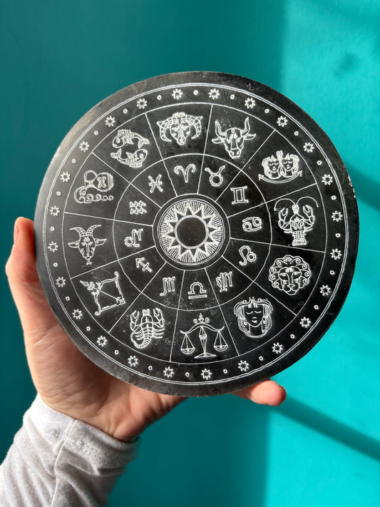 Hand holding a charging plate selenite zodiac with engraved zodiac symbols on a black circular surface against teal background