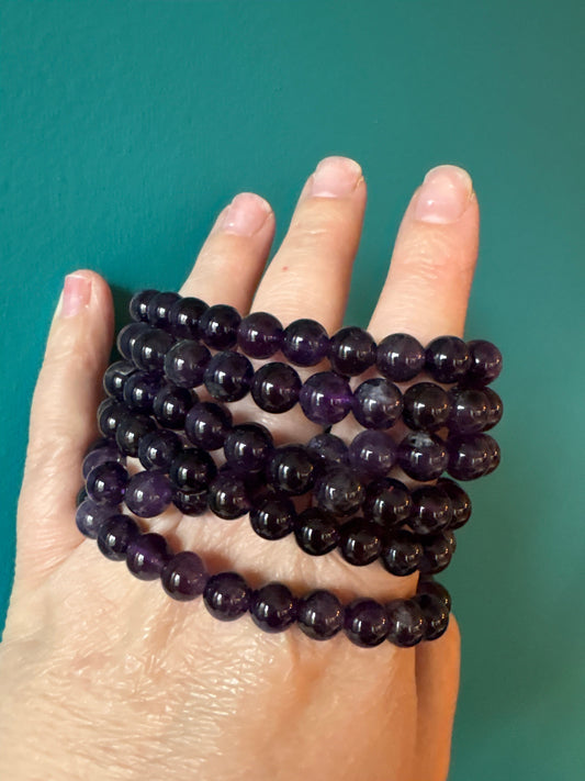 Hand holding multiple strands of polished 8mm amethyst bracelet beads against a teal background