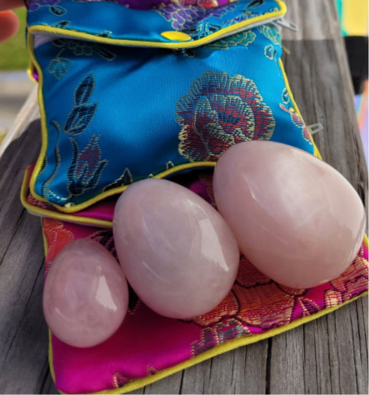 Set of three polished Rose Quartz Yoni Eggs with colorful floral pouches on wooden surface
