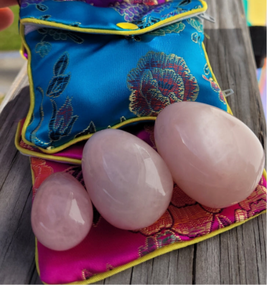 Set of three polished Rose Quartz Yoni Eggs with colorful floral pouches on wooden surface