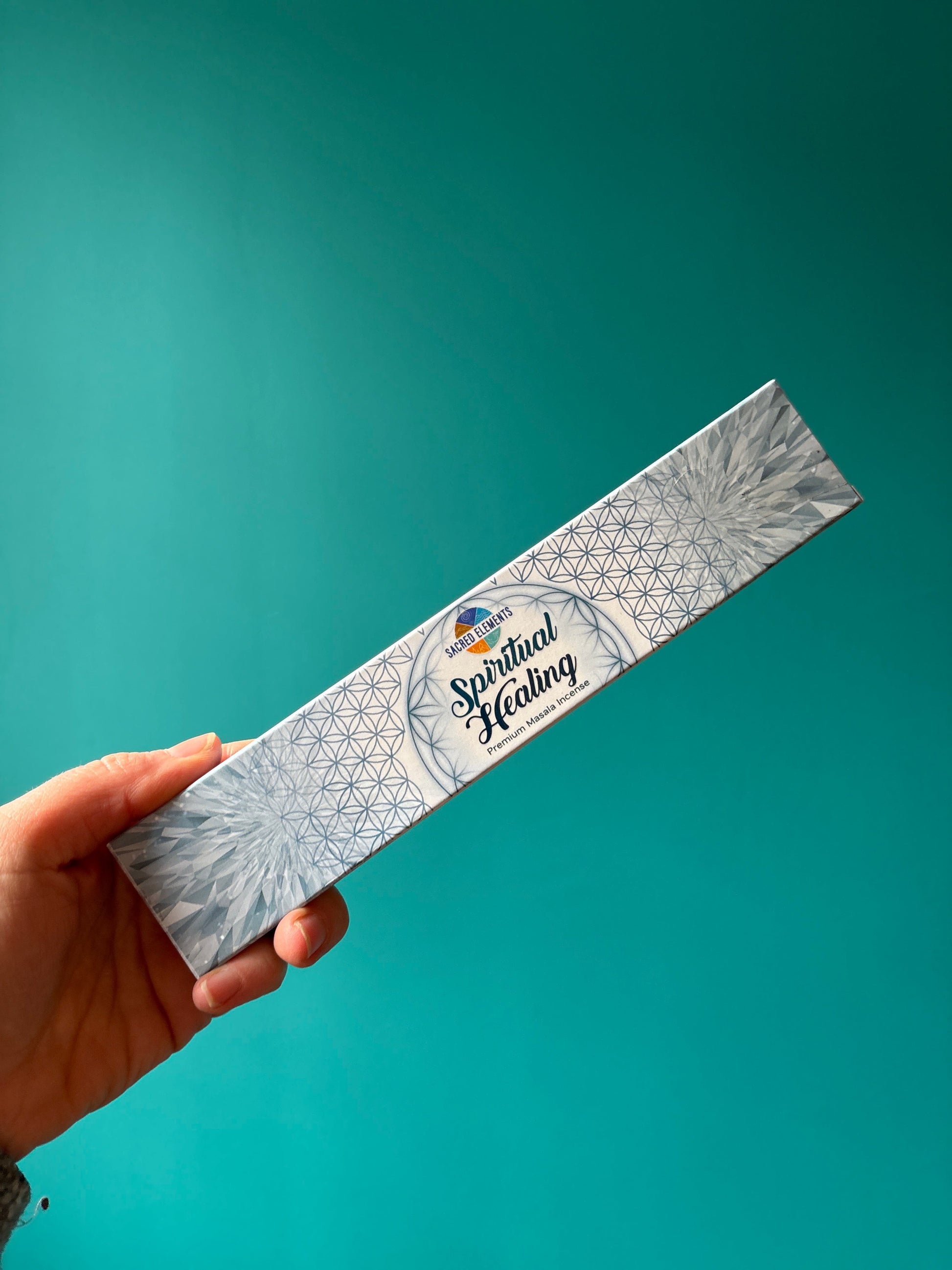 Hand holding Sacred Element Incense box with spiritual healing design against teal background