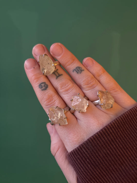 Close-up of hand wearing four rings featuring unique Libyan Desert Glass stones on a green background
