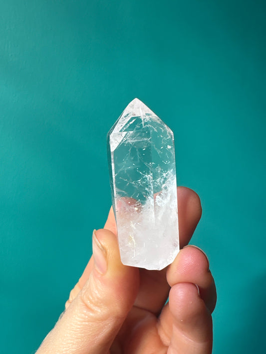 Hand holding a polished 1-2 inch clear quartz point tower crystal against a teal background