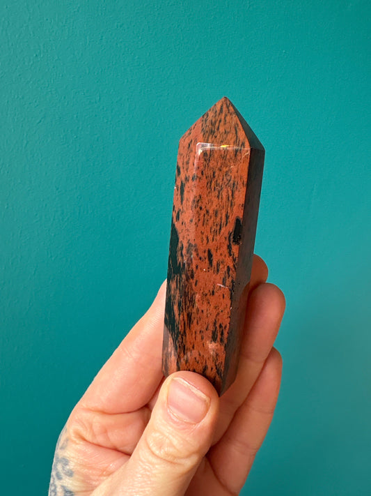Hand holding a polished mahogany obsidian tower crystal with a blue background