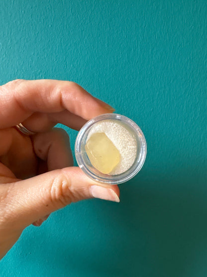 Hand holding a small container with a piece of yellow Libyan Desert Glass against a teal background