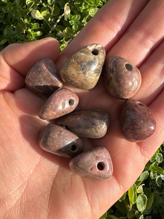 Hand holding multiple polished Rhodonite pendant stones with holes for stringing, outdoors with green foliage background