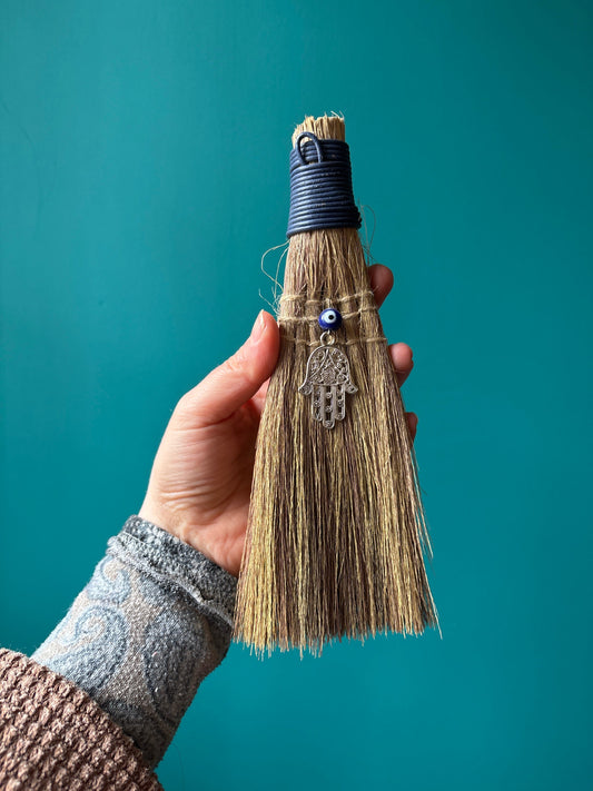 Hand holding a handcrafted witch broom with a traditional design and decorative charm against a teal background