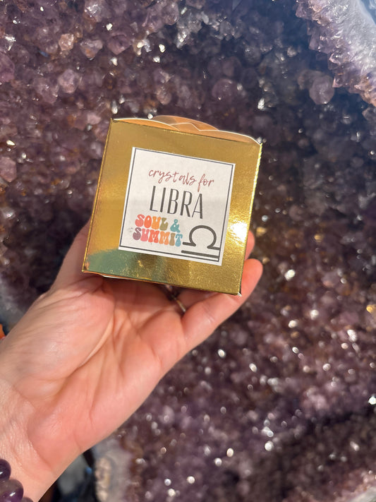 Hand holding a gold Libra Crystal Gift Box labeled "crystals for Libra" against a sparkling amethyst background