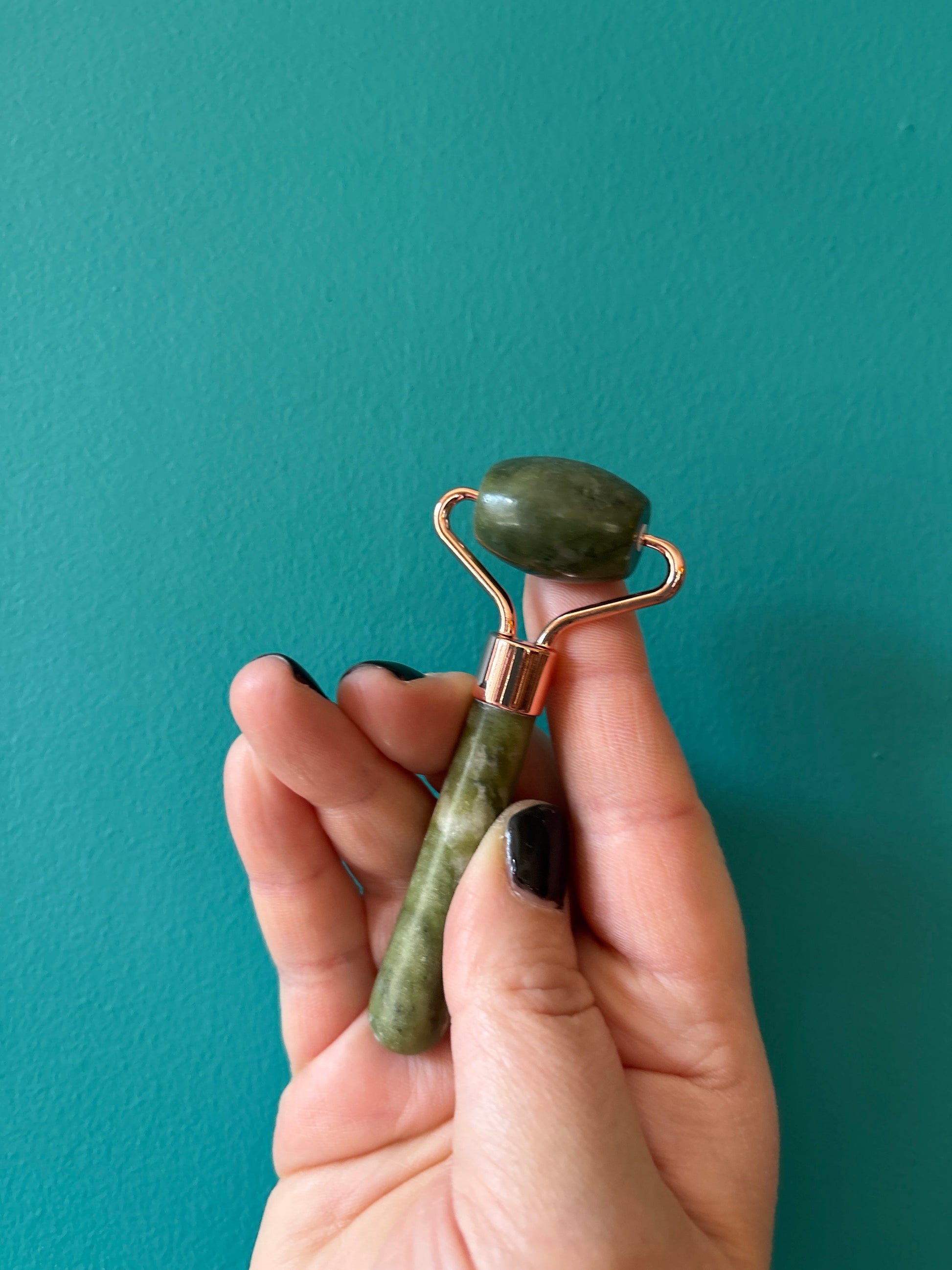 Hand holding a green jade facial massage roller against teal background