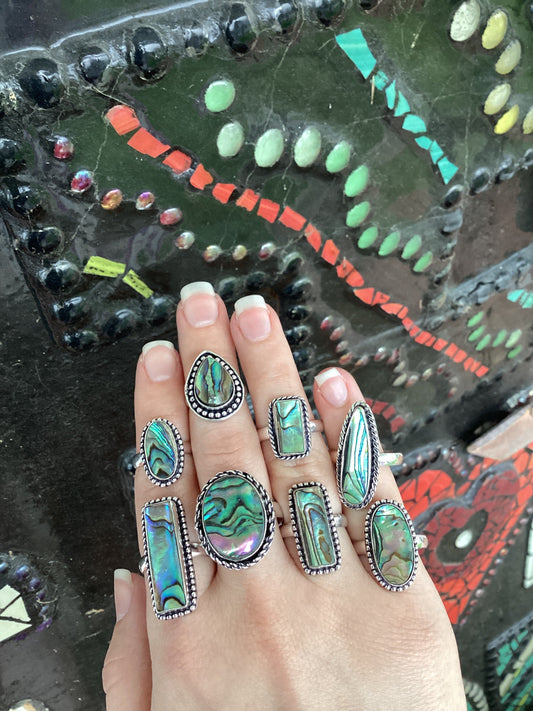 Close-up of a hand wearing multiple silver-plated abalone rings with colorful iridescent patterns.